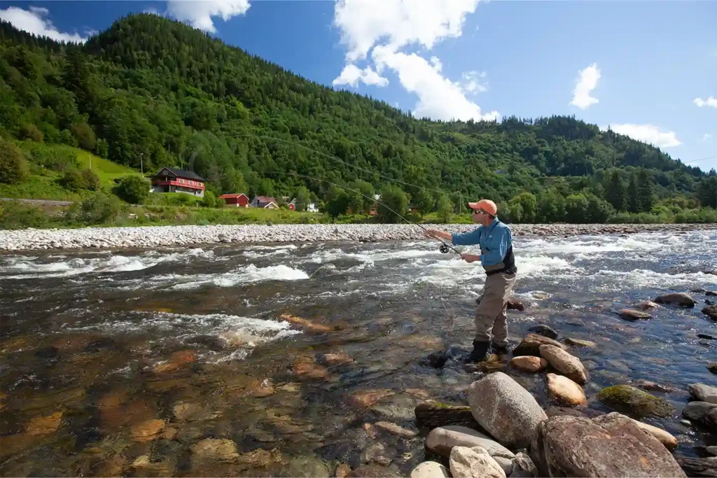 A man is fishing in a river near mountains.