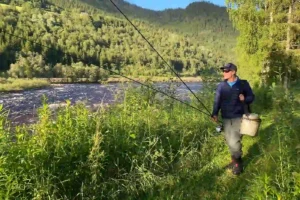 A man is standing on a grassy hill next to a river, fly fishing.