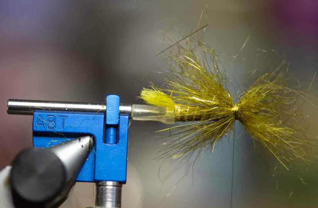 A close-up of a fishing fly being tied in a vise, with gold and yellow fibers creating the body and wings.