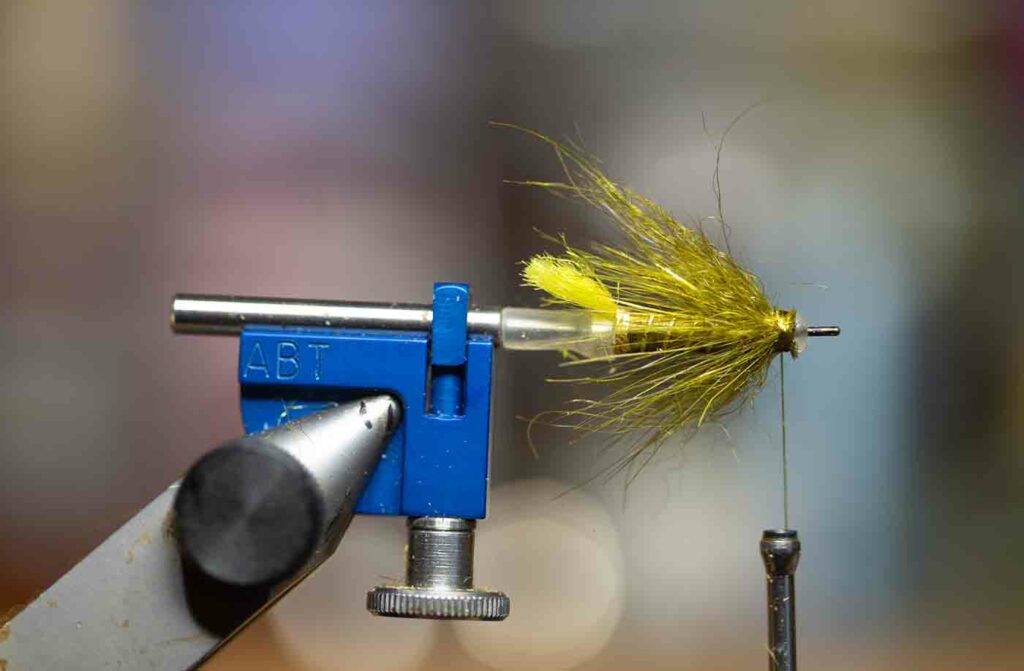 Close-up of a fly tying vise holding a partially completed artificial fishing fly with yellow and green materials attached.
