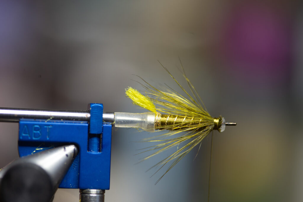 A close-up of a yellow artificial fishing fly held in a blue vise, used for fly tying.