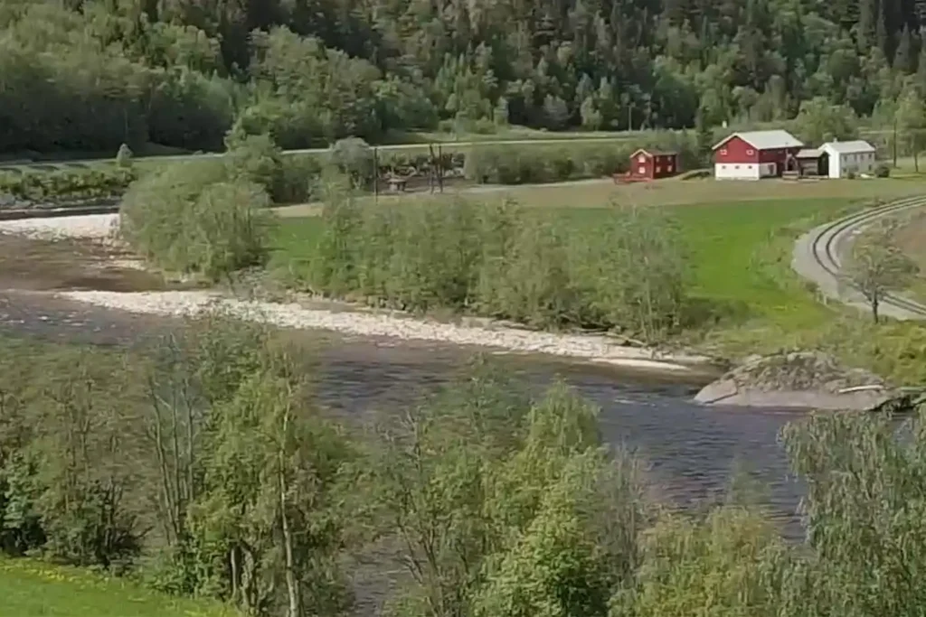 A river flows through a green landscape with trees, a road, and several red and white buildings visible in the background.